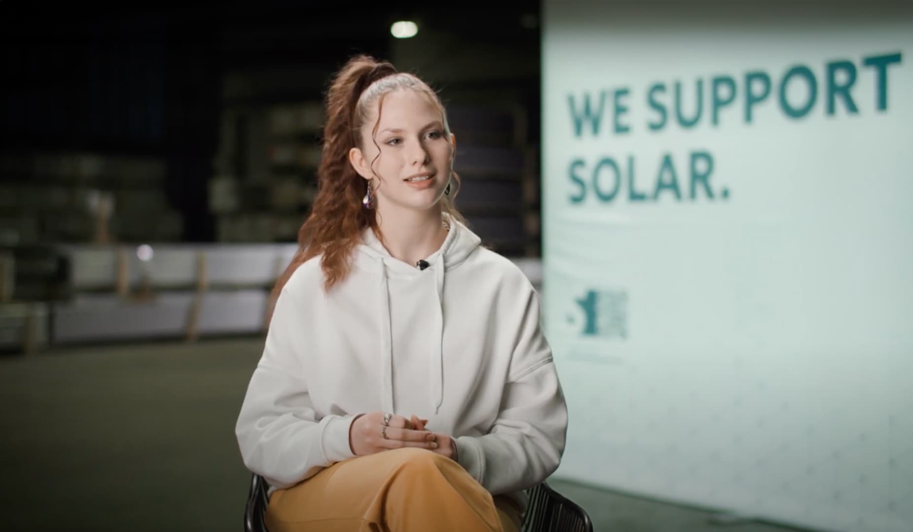 A woman with a ponytail sits in a chair, speaking. Behind her is a banner that reads, "We support solar." She is wearing a white hoodie and yellow pants.