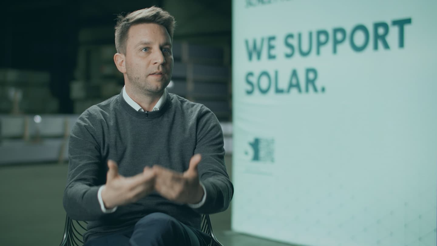 A man is seated indoors, gesturing with his hands. Behind him is a sign that reads "WE SUPPORT SOLAR.