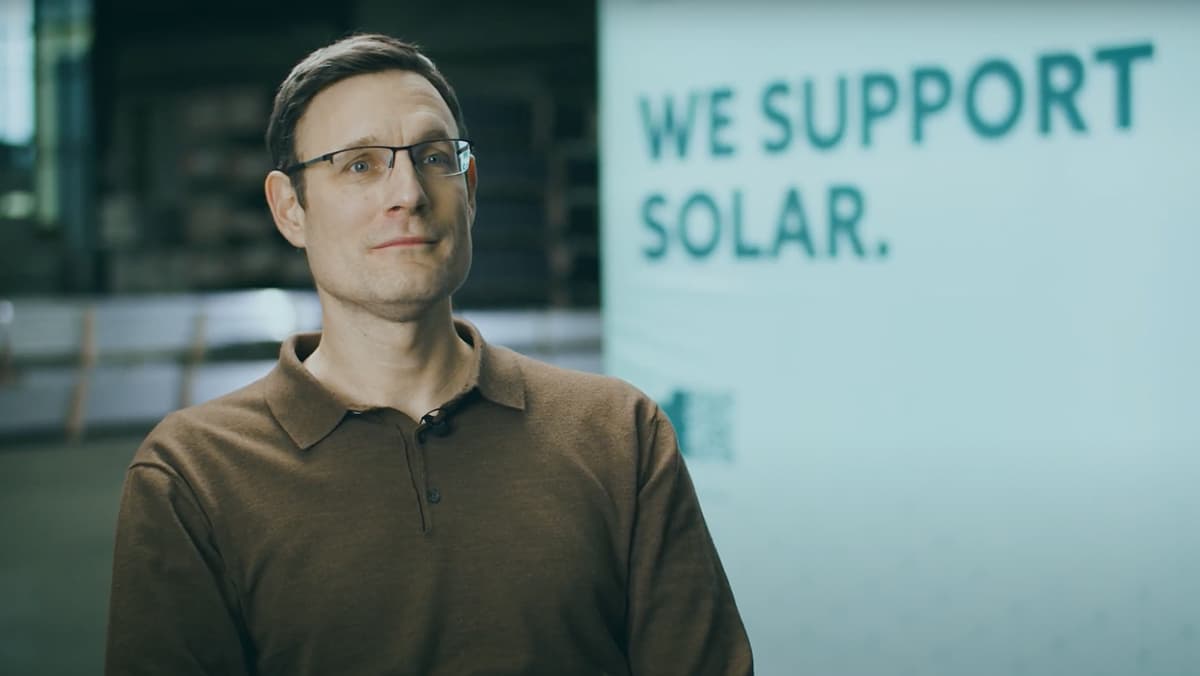 A man wearing glasses and a brown shirt stands in front of a sign that reads, "We Support Solar.