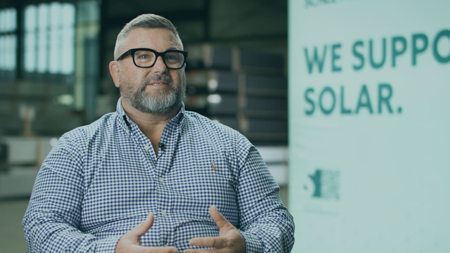 A man with glasses and a beard is speaking, sitting in front of a sign that partially reads "WE SUPPORT SOLAR.