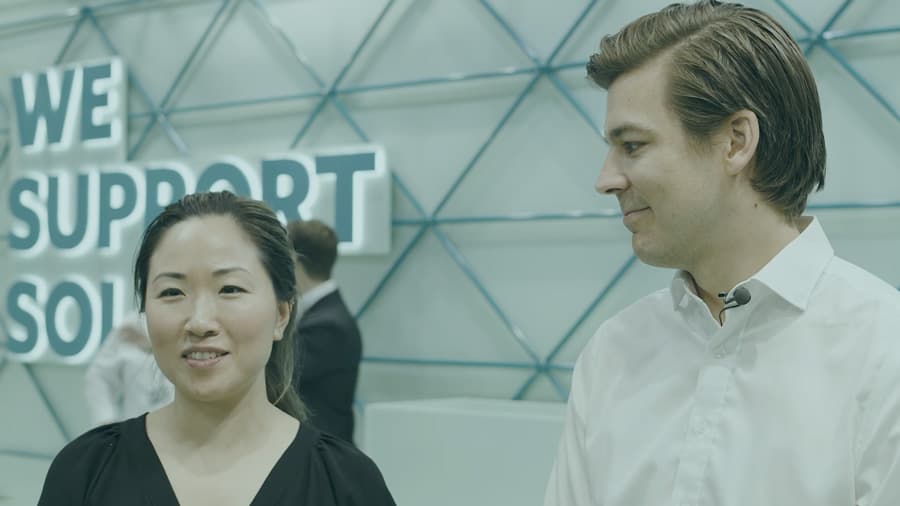 Two people, one wearing a black shirt and the other wearing a white shirt, stand in front of a sign that reads "We Support Sol".