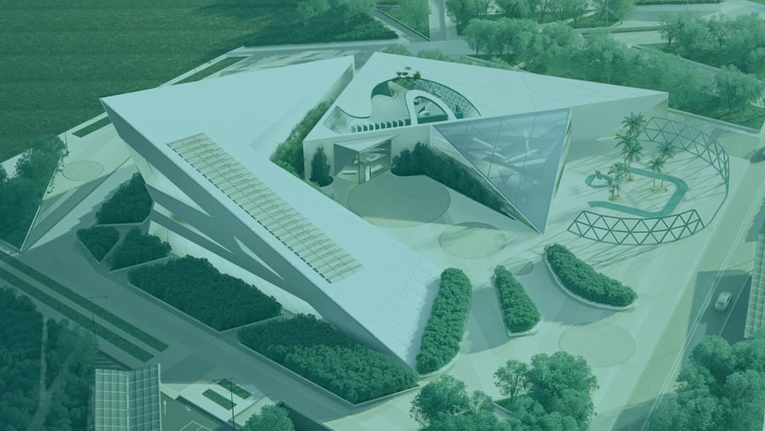 Aerial view of a large, modern, angular building with geometric shapes, green spaces, and a glass dome roof. Surrounded by trees and a road on the right side.