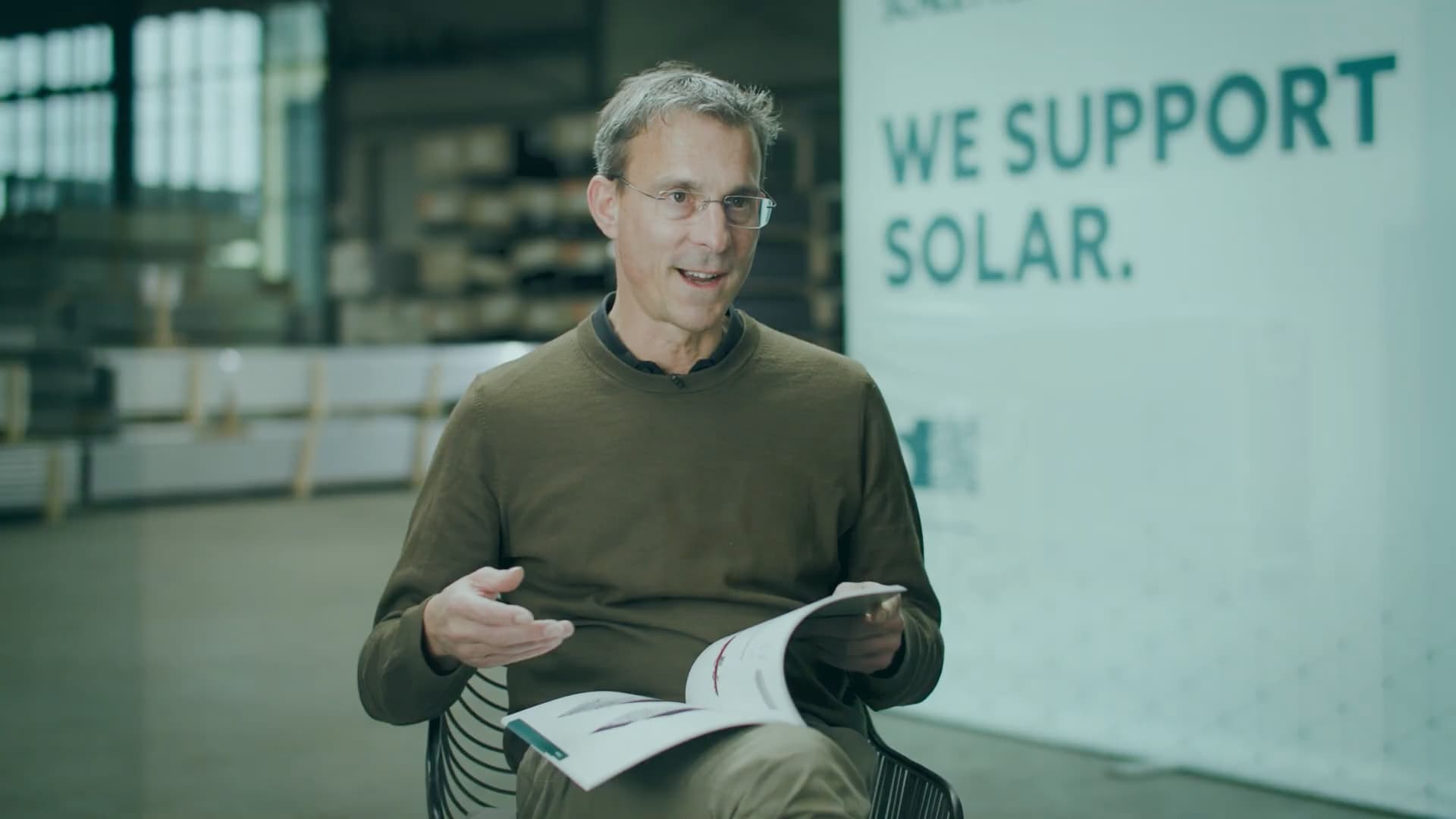 A seated man holds a brochure and speaks in front of a blurred backdrop with the text "We Support Solar.