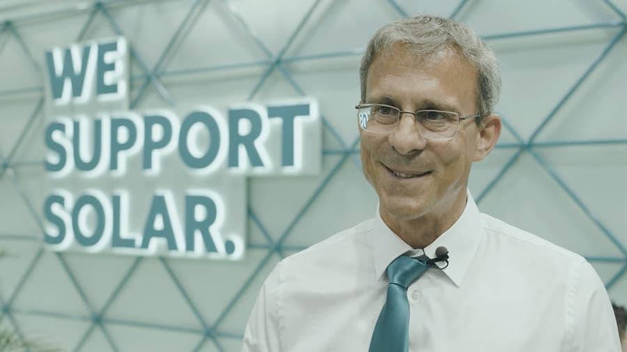 A man in a white shirt and blue tie stands smiling in front of a wall with the words "We Support Solar" displayed.