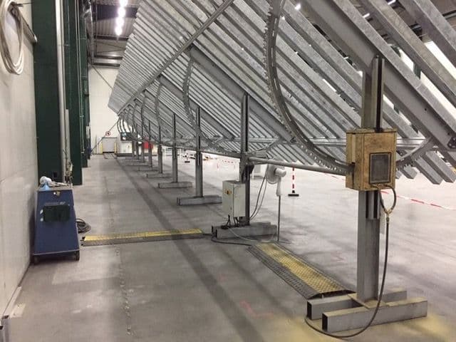 A long, industrial metal structure with numerous angled supports extending the length of a large warehouse-like facility, with various control equipment and a ventilation fan nearby.