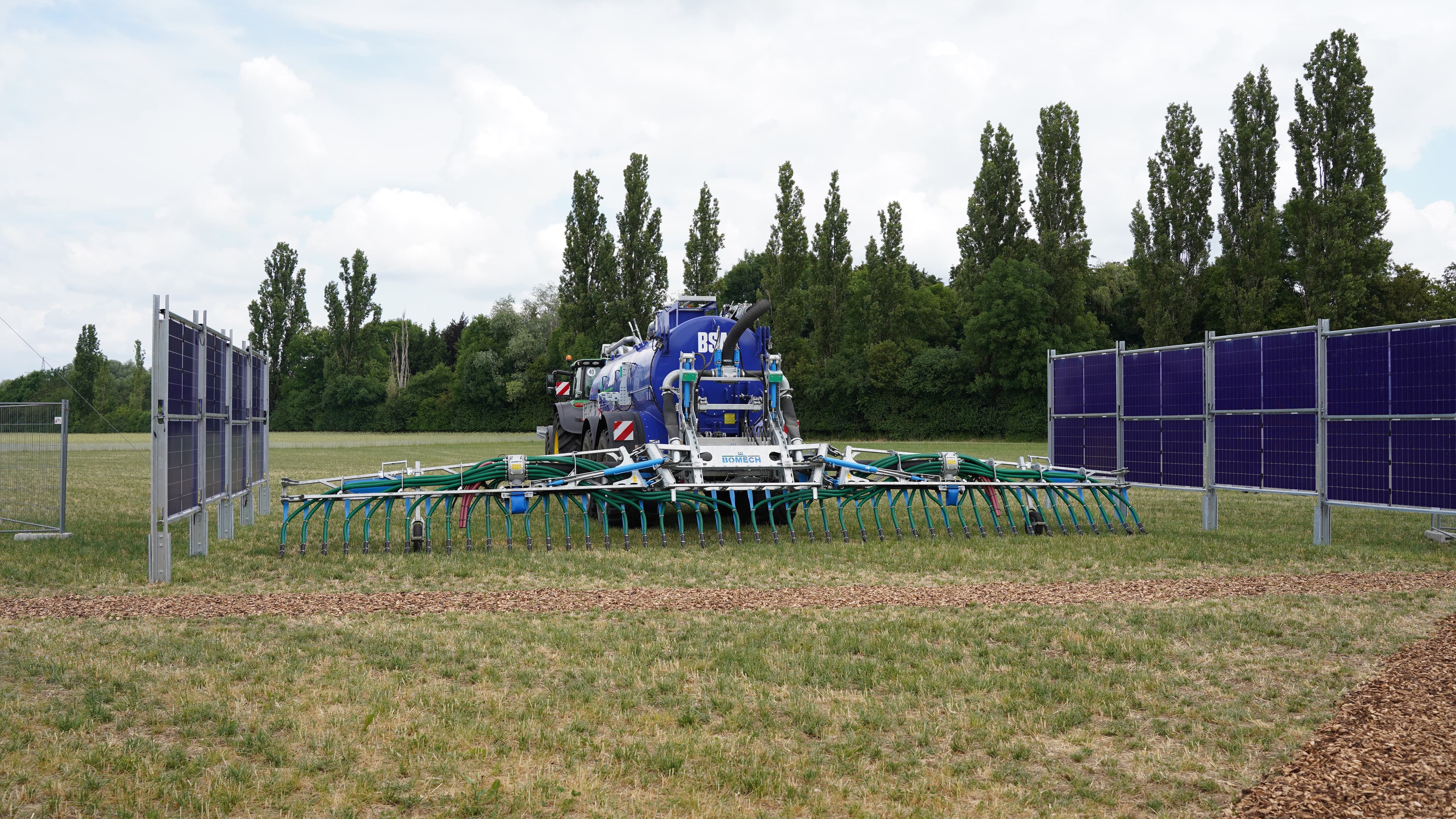 A large agricultural machine operates between rows of solar panels in a grassy field with trees in the background.