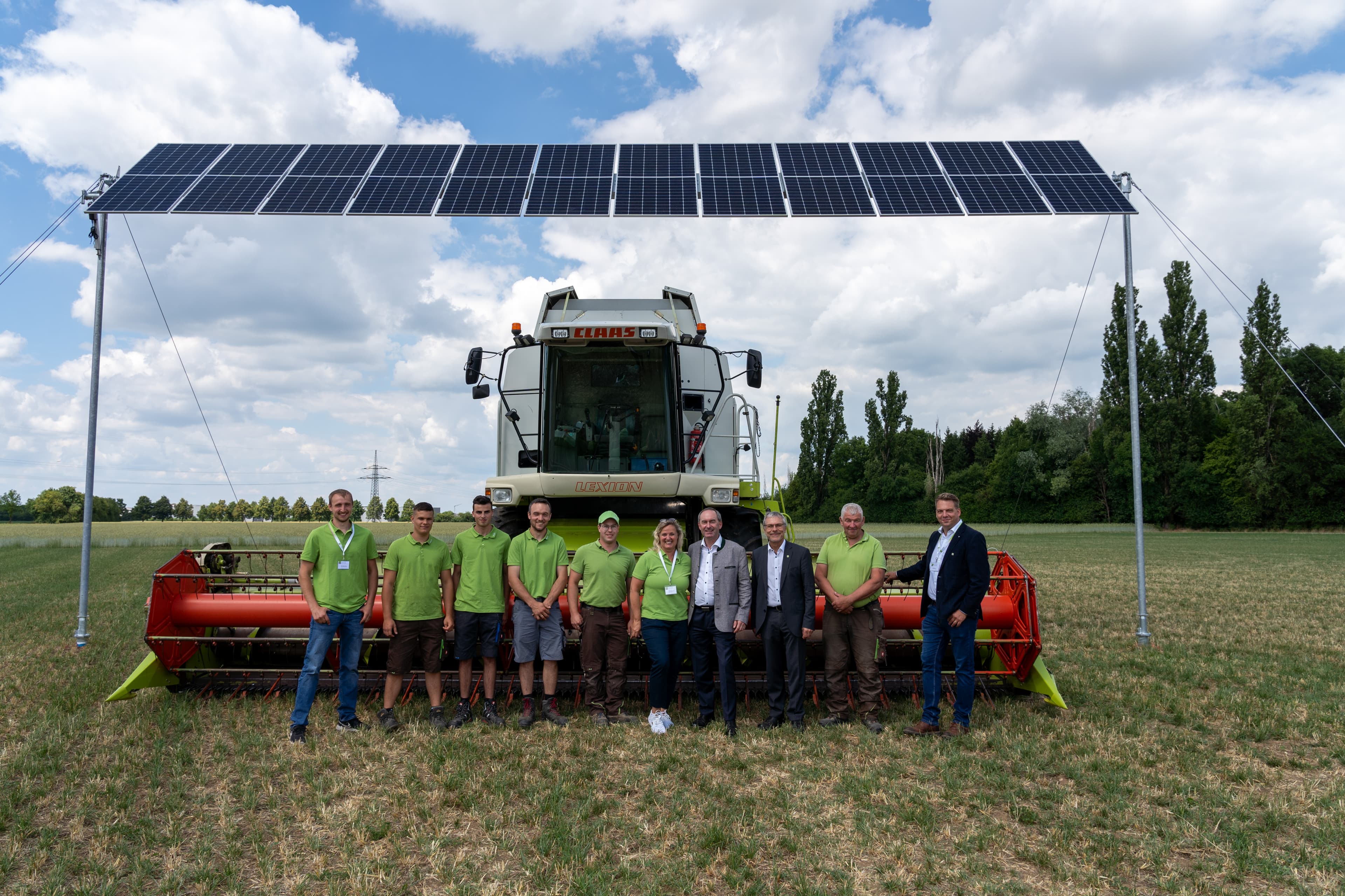 Group of people standing in front of a large agricultural machine under a solar panel structure in a field with a partly cloudy sky in the background.