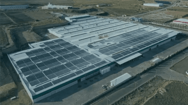 A large industrial building with extensive solar panel installations on the roof, surrounded by a barren landscape with some roads and additional facilities in the distance.