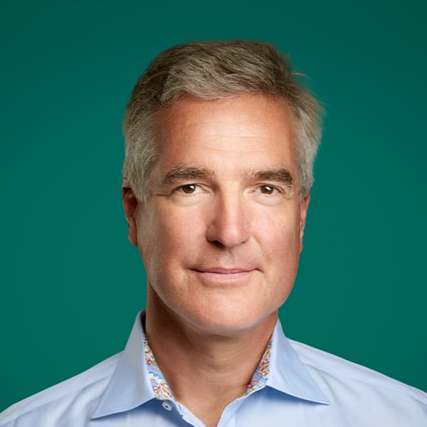A man with gray hair wearing a light blue collared shirt against a green background.
