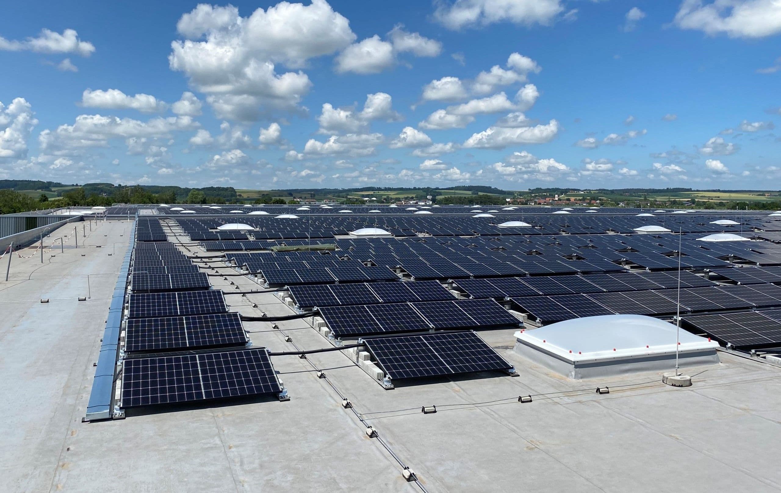 Solar panels covering a large rooftop under a sky with scattered clouds. The panels are arranged in rows, and the landscape is visible in the background.