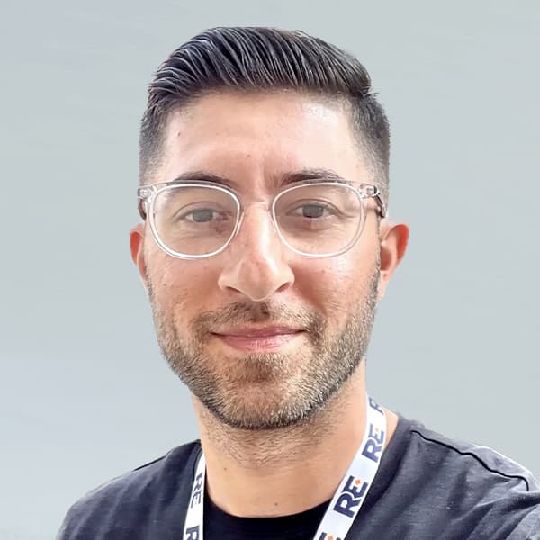 A person with short, dark hair and glasses is wearing a black shirt and lanyard. They are smiling and posing against a gray background.