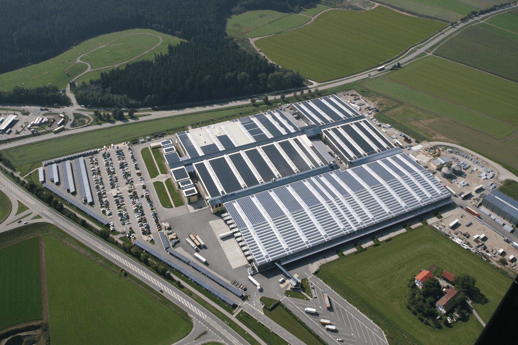 Aerial view of a large industrial facility with multiple buildings, parking areas, and surrounding fields. A highway and roads are visible nearby.