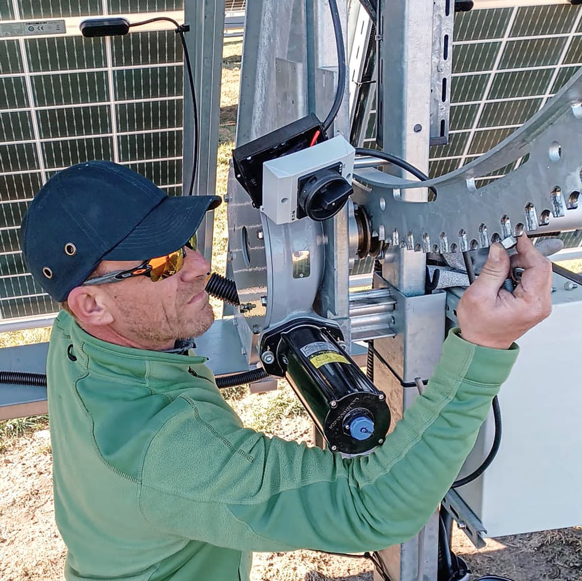 A person in a green jacket and black cap adjusts equipment on a solar panel structure.