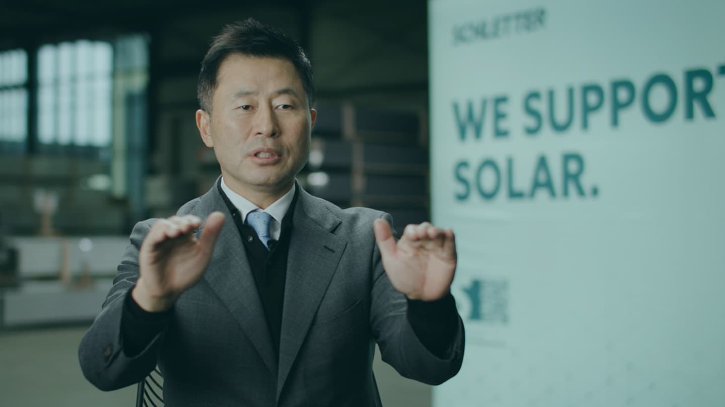 A man in a suit and tie is speaking, gesturing with both hands. A banner in the background reads "WE SUPPORT SOLAR.