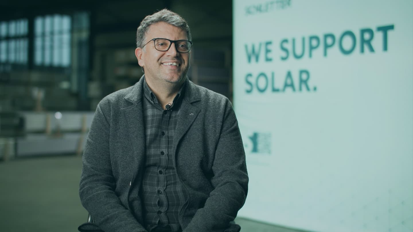 A man wearing glasses and a gray blazer sits in front of a sign that reads, "We support solar." The background is an indoor setting.