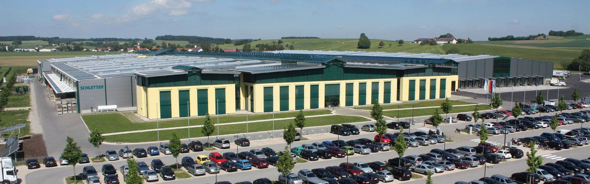 A wide aerial view of a large industrial building with multiple joined sections, surrounded by a full parking lot, set in a rural area with fields and distant houses.