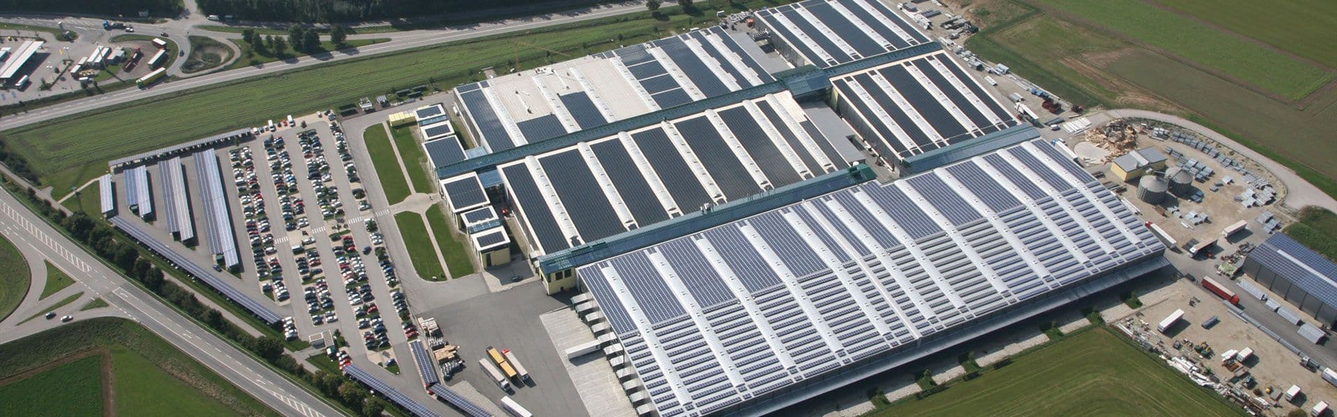 Aerial view of a large industrial facility with multiple rectangular buildings, parking lots, and surrounding green fields.