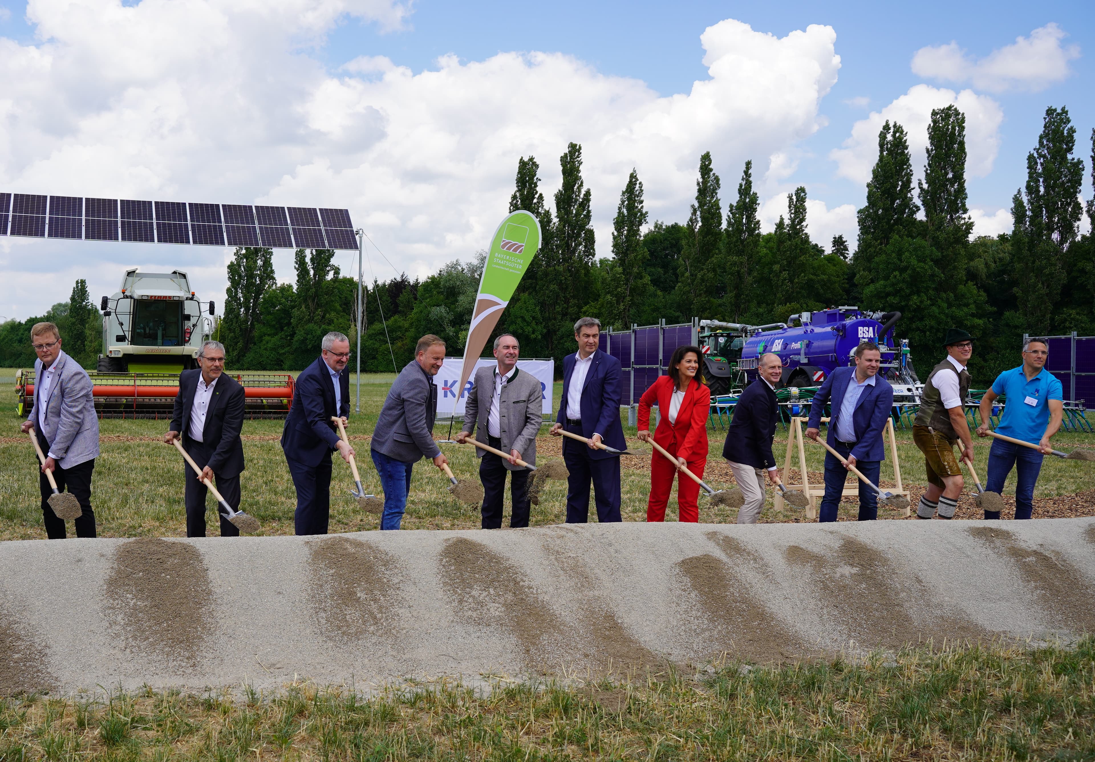 A group of people in business and casual attire participate in a groundbreaking ceremony at a construction site, holding shovels and standing in front of a mound of dirt with machinery and greenery in the background.