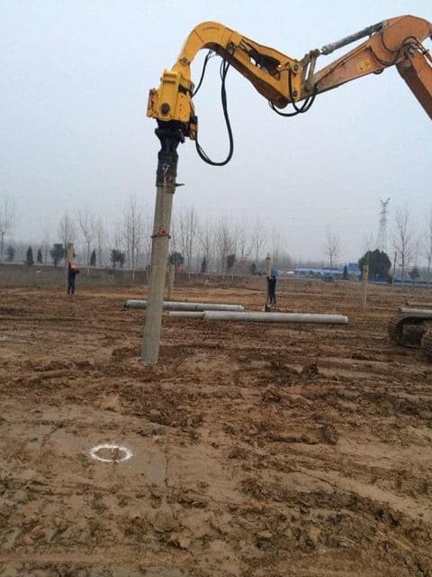A construction site with an excavator placing a pole into the ground. Two workers stand in the background. The ground is muddy, and there are trees and pipes around.