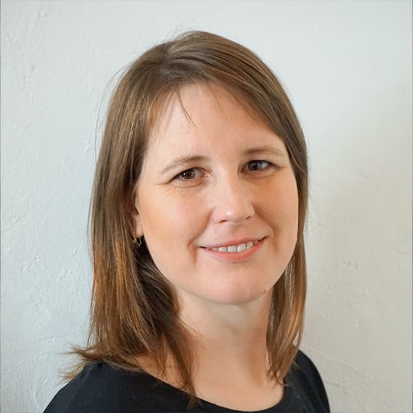 A woman with shoulder-length brown hair and a black top smiles in front of a plain light-colored background.
