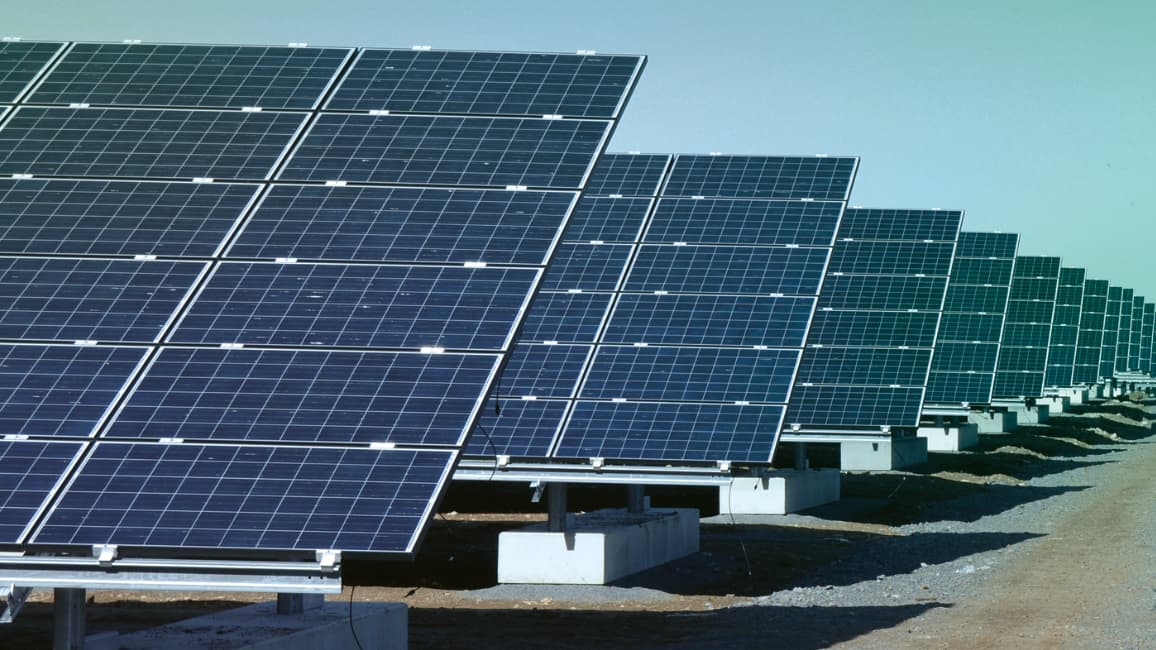 Rows of large solar panels are installed on the ground in an open area, capturing sunlight for renewable energy. The sky is clear.