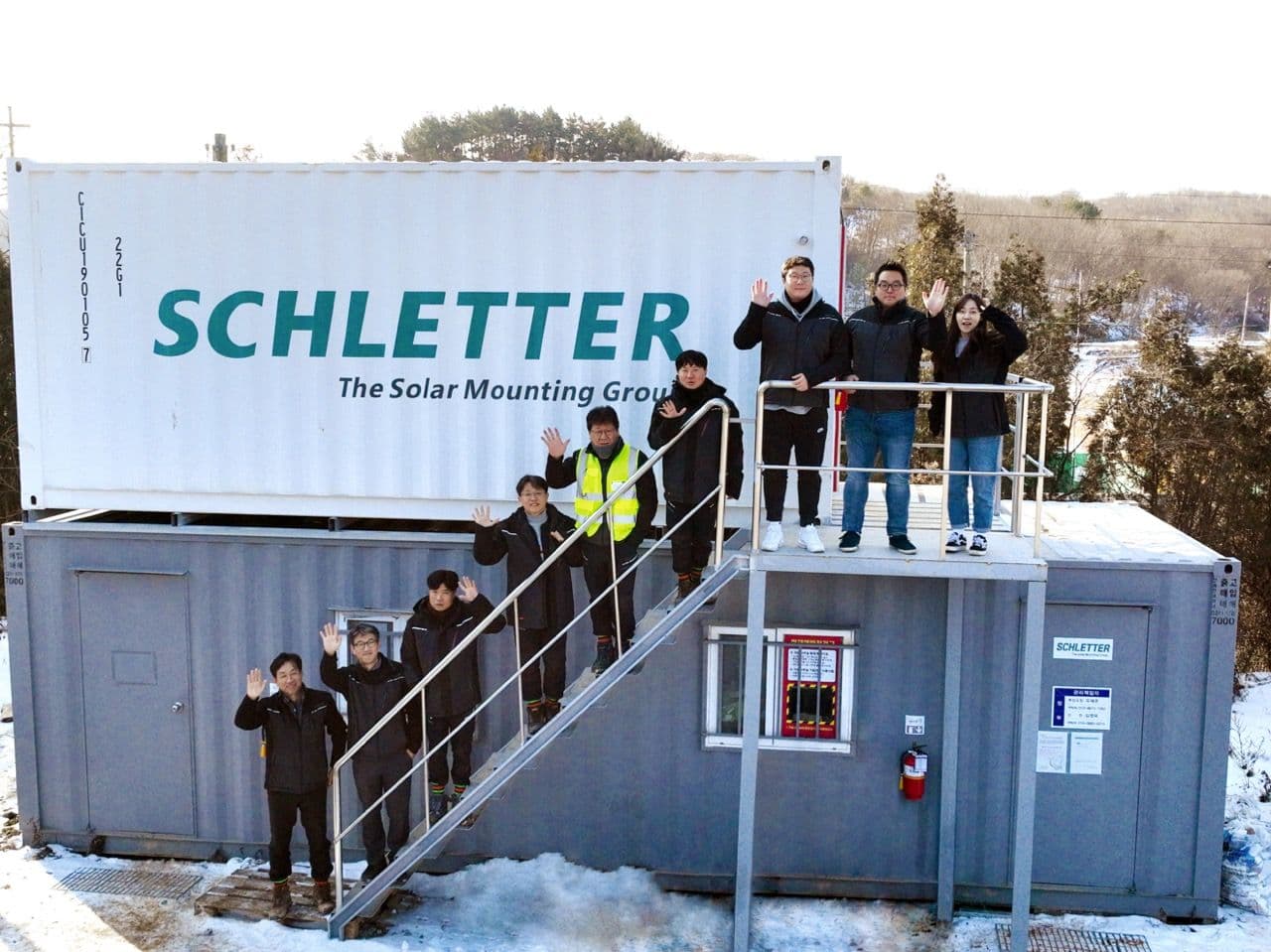 A group of people stand on and next to stairs leading to a shipping container with "Schletter, The Solar Mounting Group" written on it. Snow is on the ground, and they are waving at the camera.