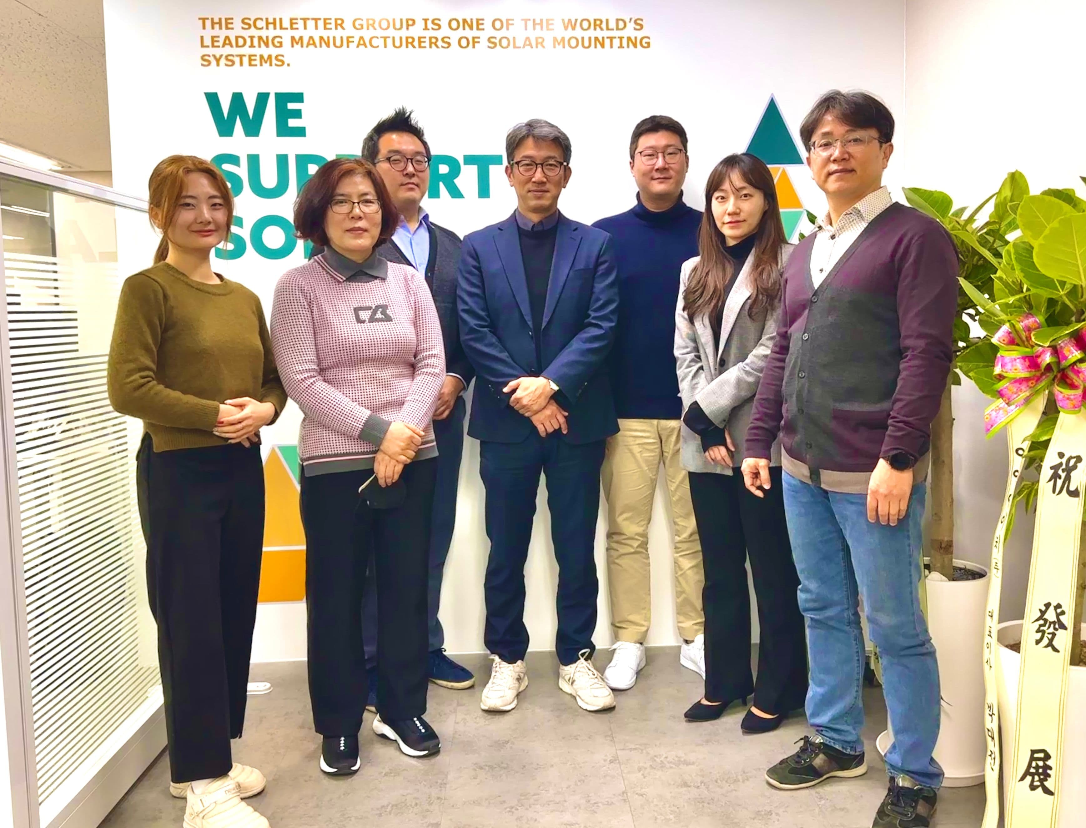 Seven people are standing indoors in front of a sign for Schletter Group, a manufacturer of solar mounting systems.