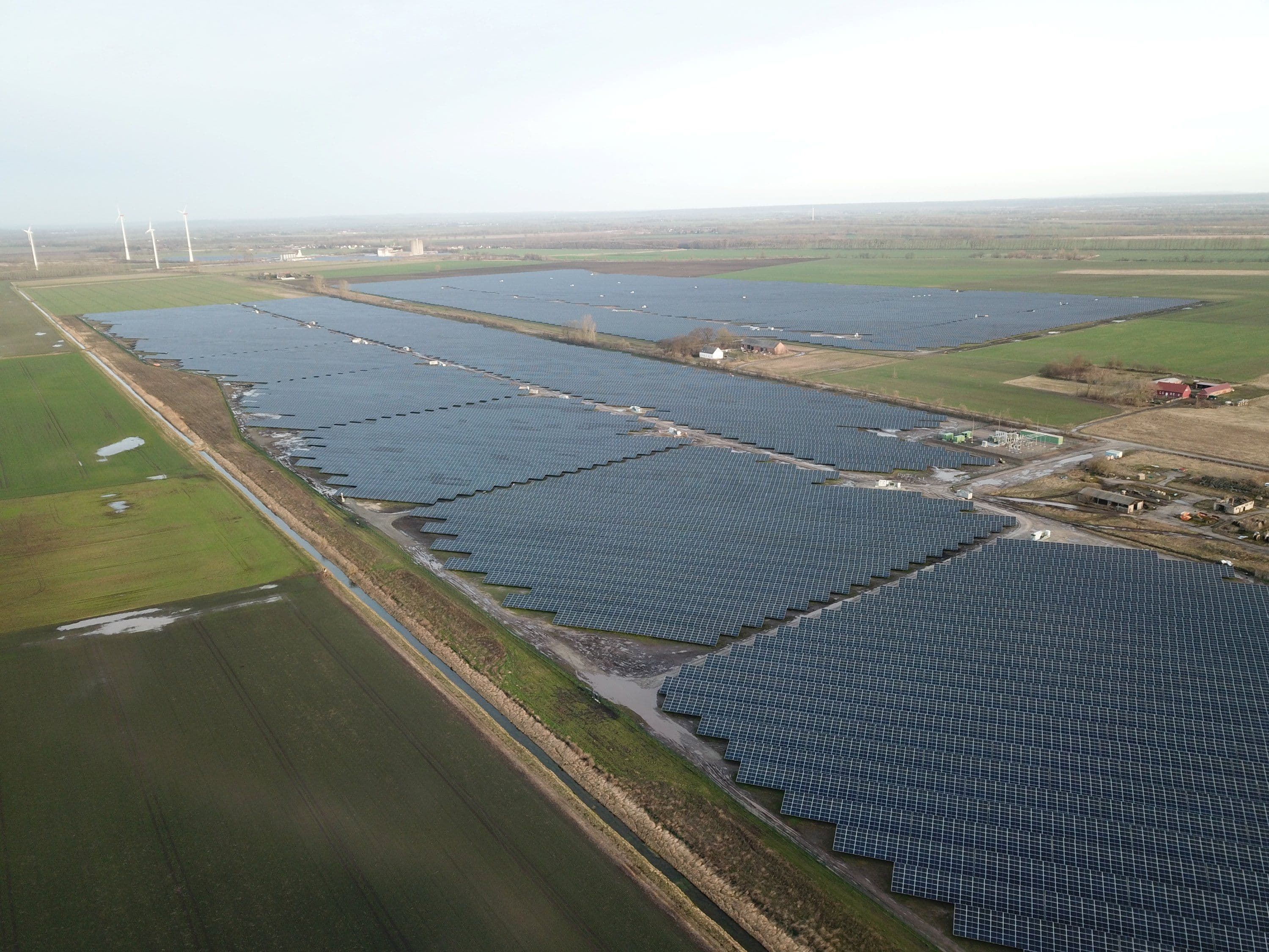 A large solar farm in a flat, rural landscape with multiple rows of solar panels stretching into the distance. The surrounding area consists of green fields and a few scattered structures.