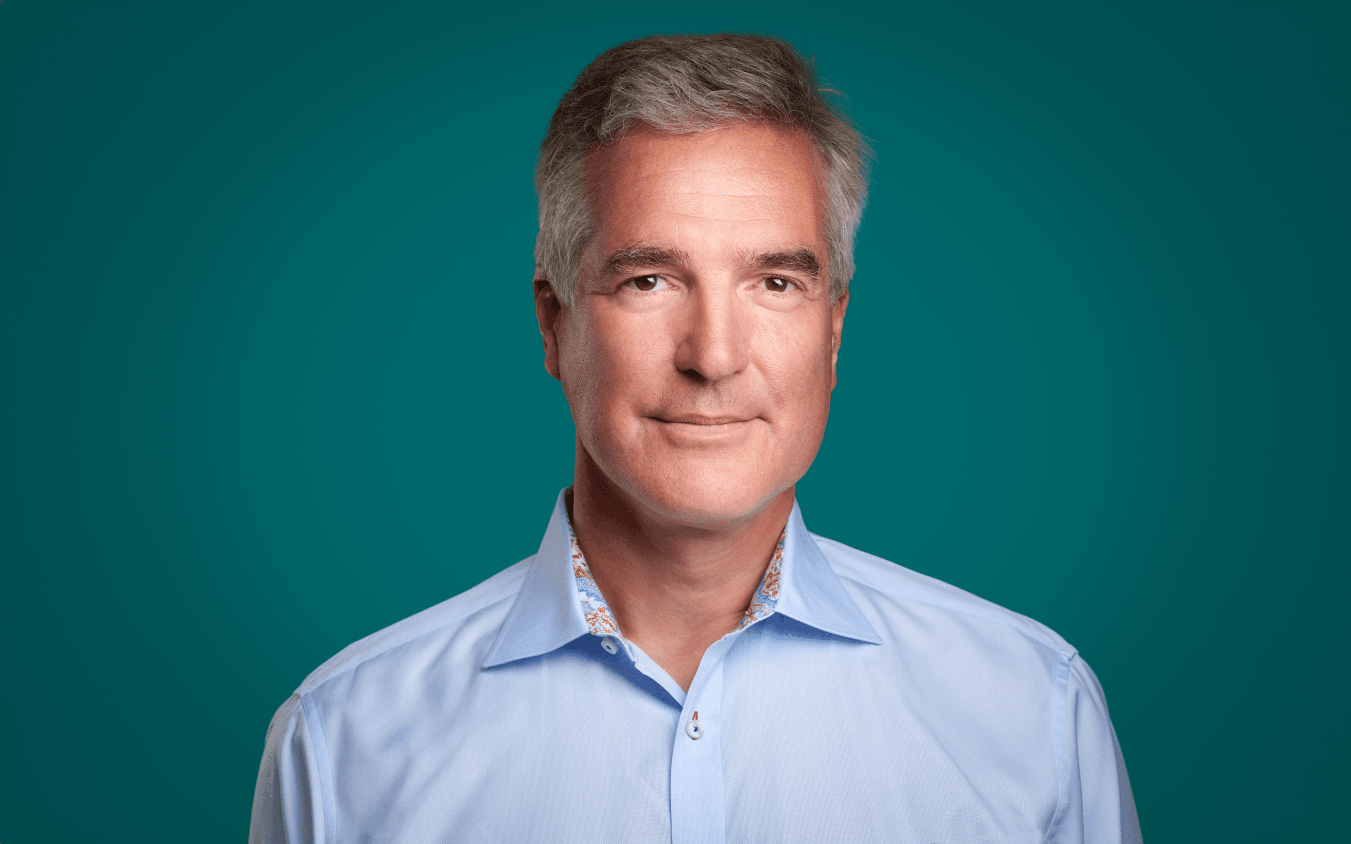 A man with gray hair is wearing a light blue collared shirt and standing against a teal background while looking at the camera.