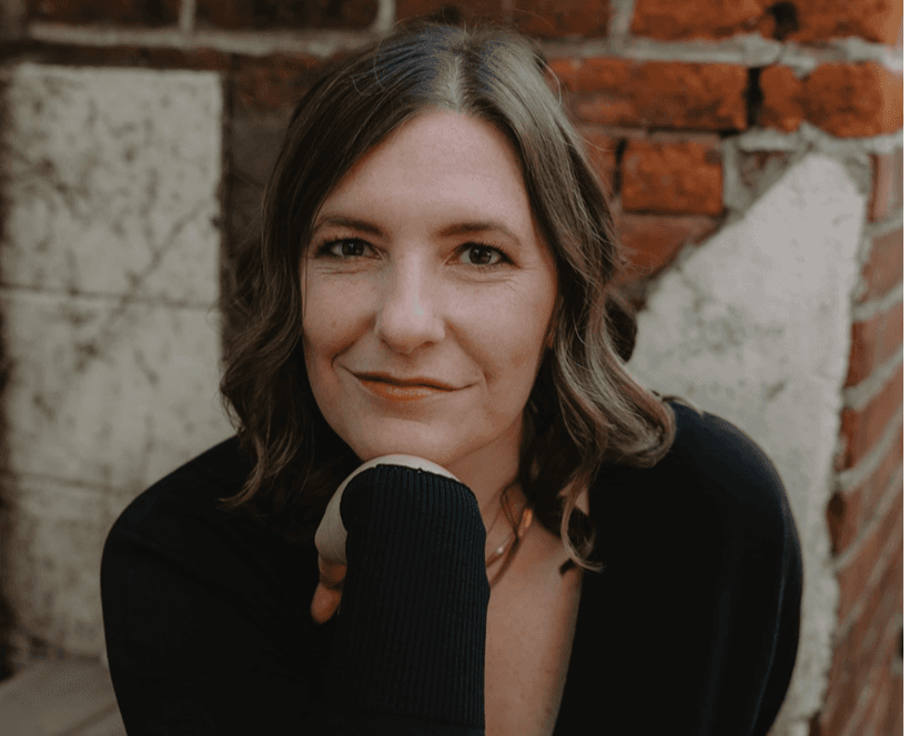 A person with shoulder-length hair and a slight smile rests their chin on their hand, posing against a brick wall.