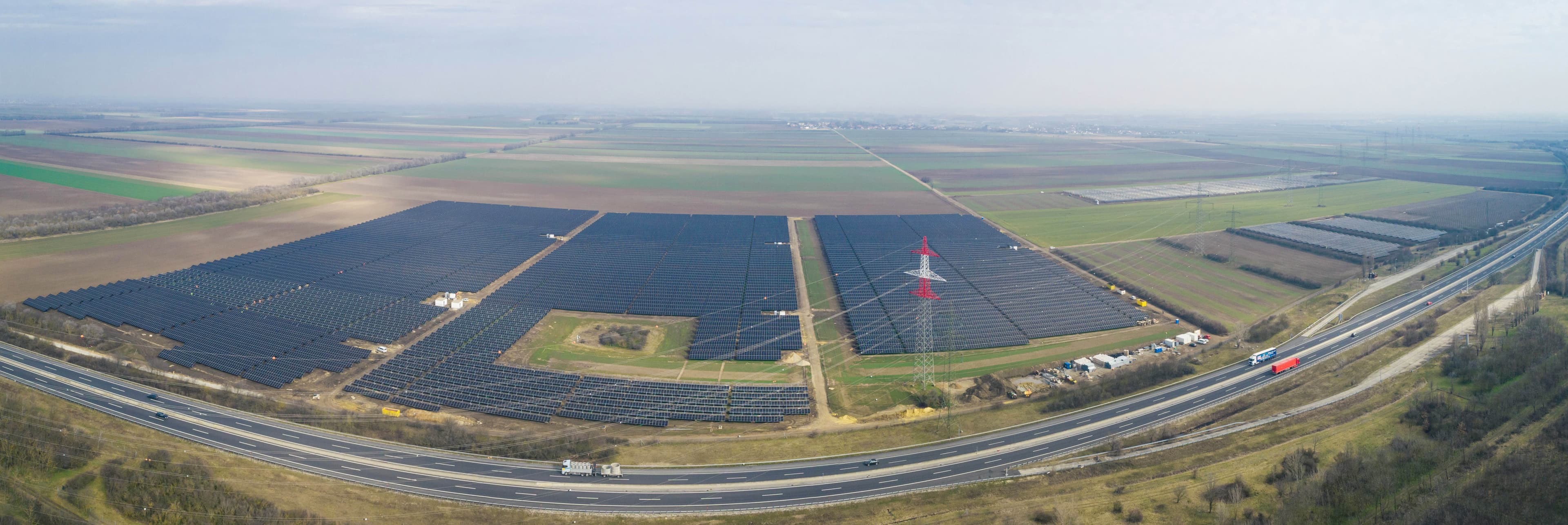 Aerial view of a large solar farm adjacent to a highway with fields and a few buildings in the background.