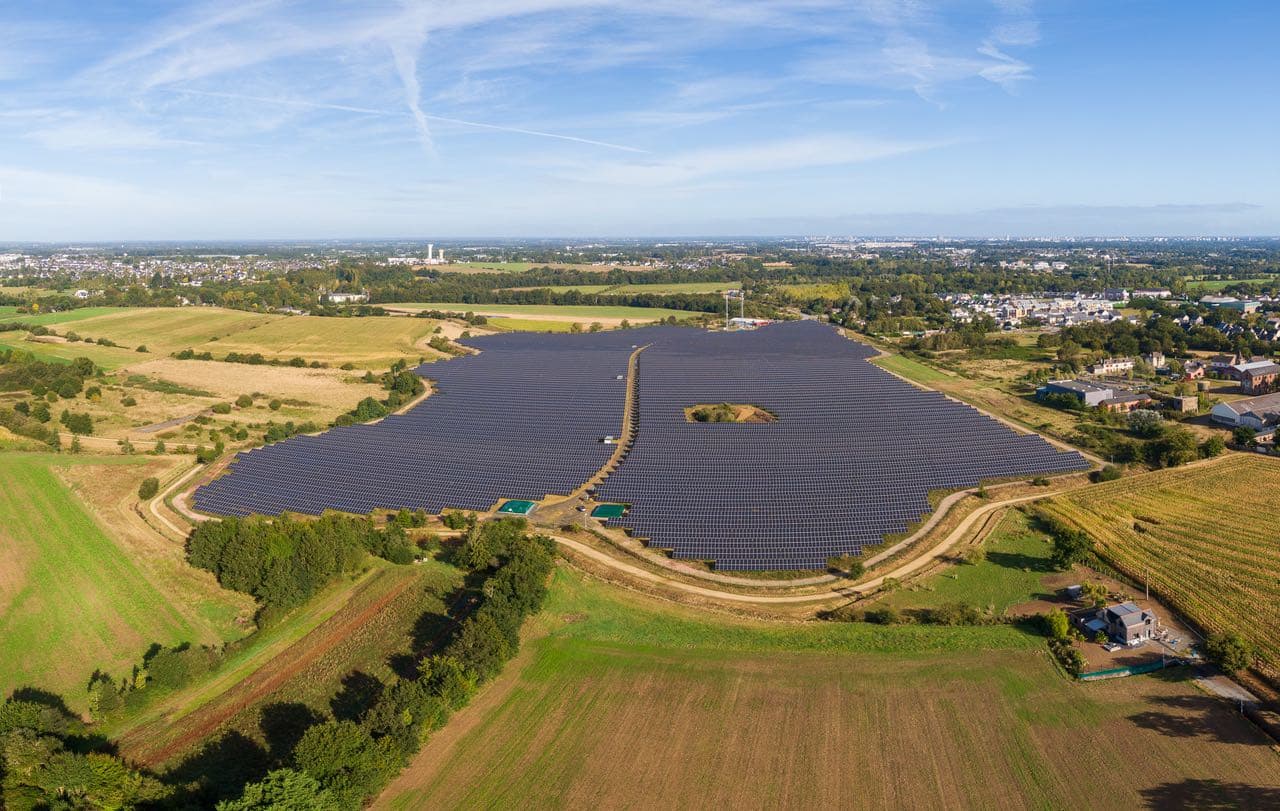 Aerial view of a large solar farm with rows of solar panels surrounded by fields and greenery, with a town visible in the distance under a blue sky.