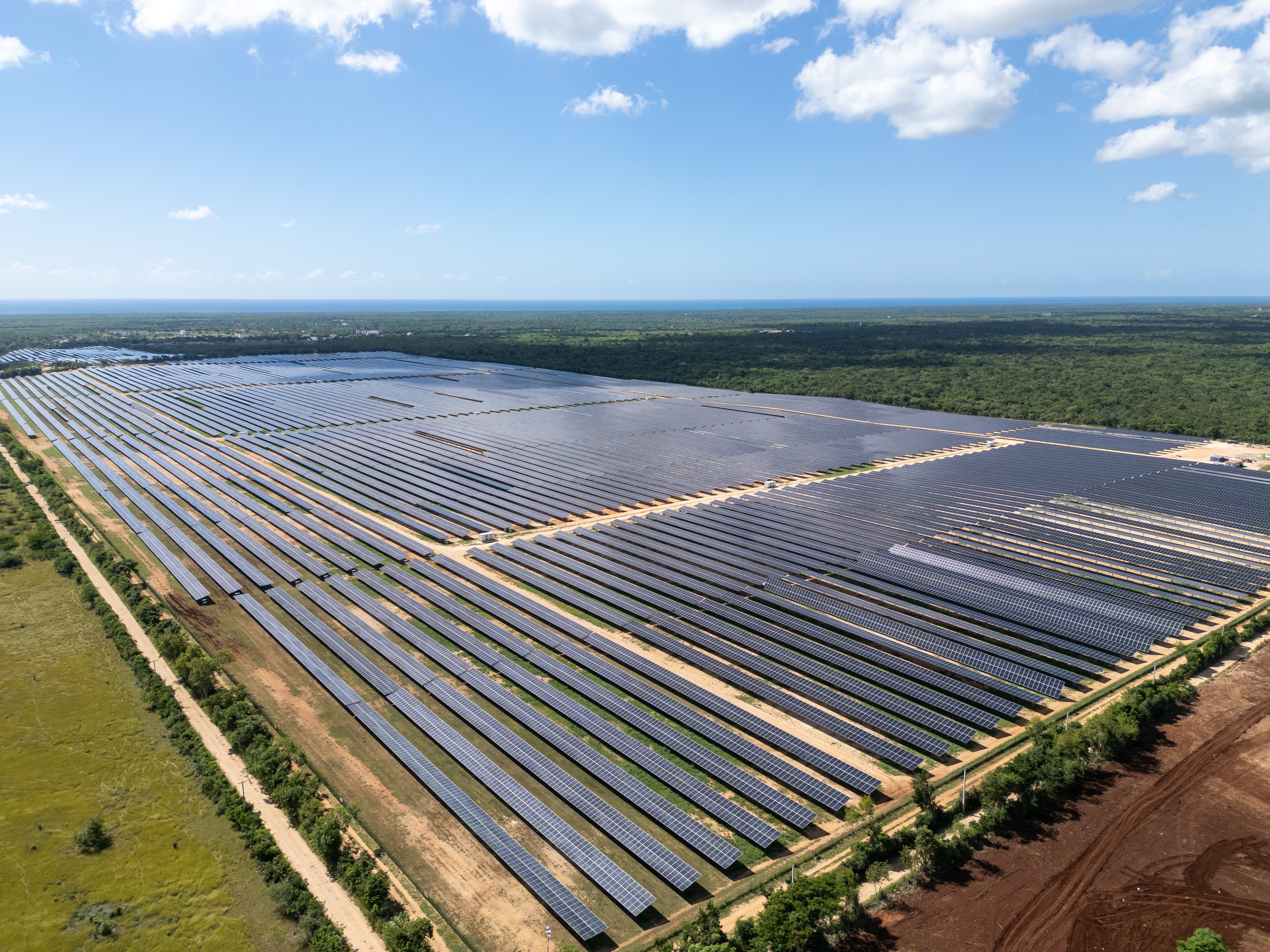 Aerial view of an expansive solar farm with rows of solar panels stretching into the distance against a backdrop of green fields and a partly cloudy sky.