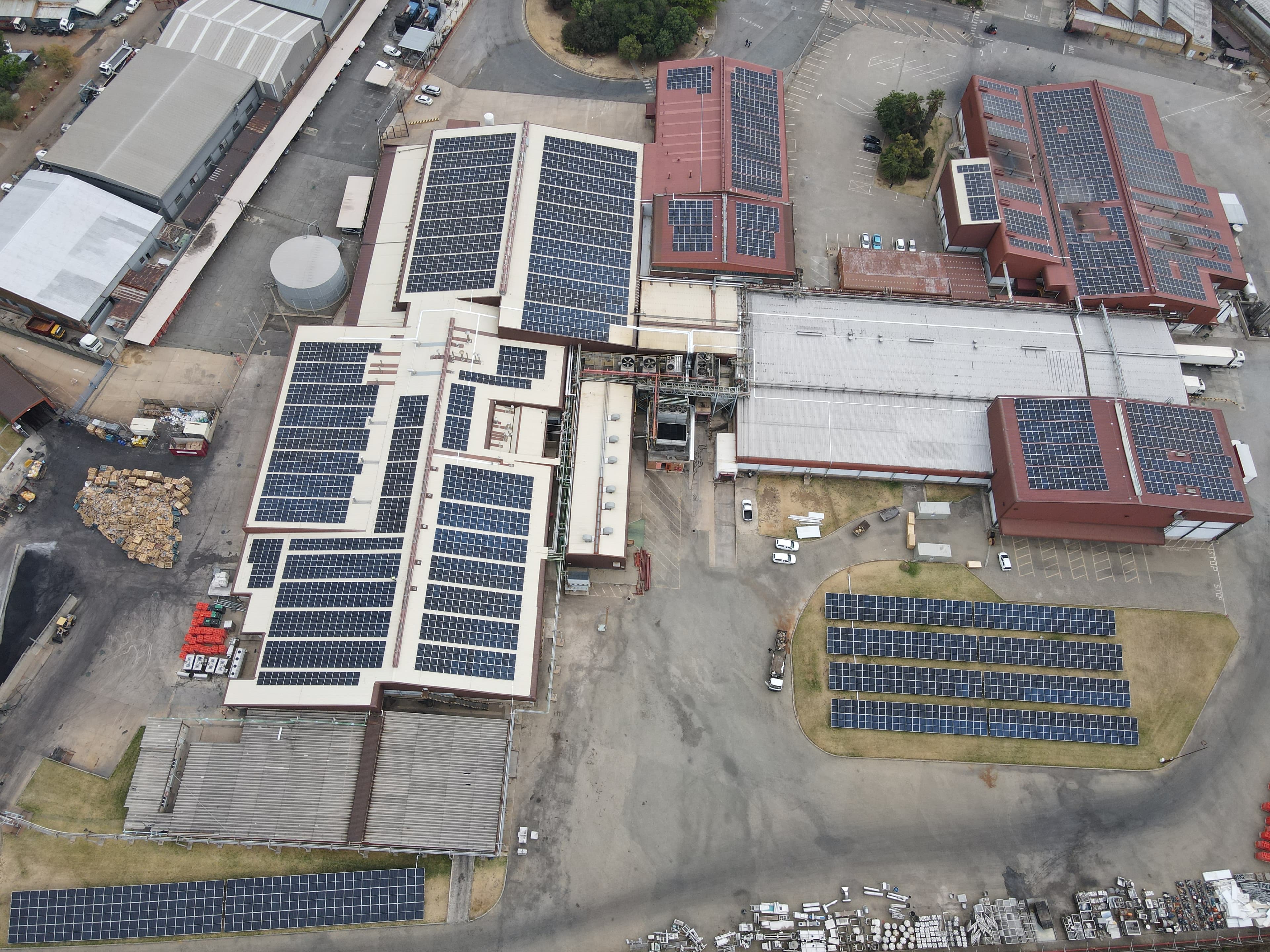 Aerial view of an industrial complex featuring multiple buildings with solar panels installed on their rooftops and additional solar panels positioned on the ground.