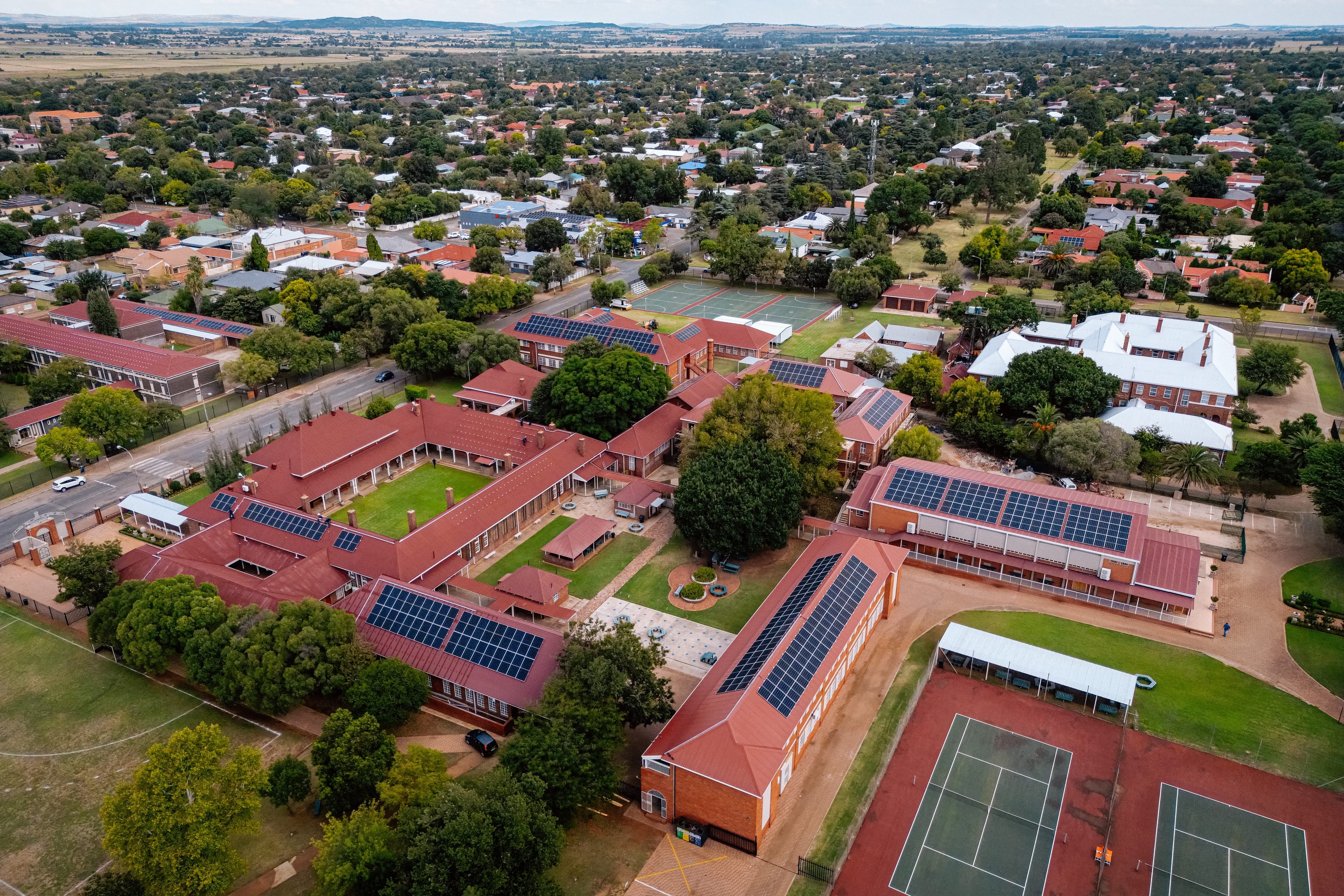 Aerial view of a large school campus with multiple red-roofed buildings, tennis courts, and surrounding trees in a suburban area. Solar panels are mounted on many rooftops.