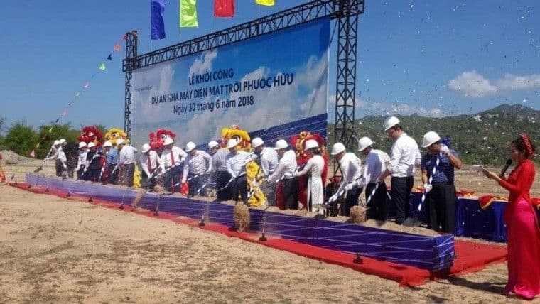 A group of people in business attire and hard hats participate in a groundbreaking ceremony for the Phuoc Huu solar power plant project, with a sign and decorated shovels in front of a large banner.