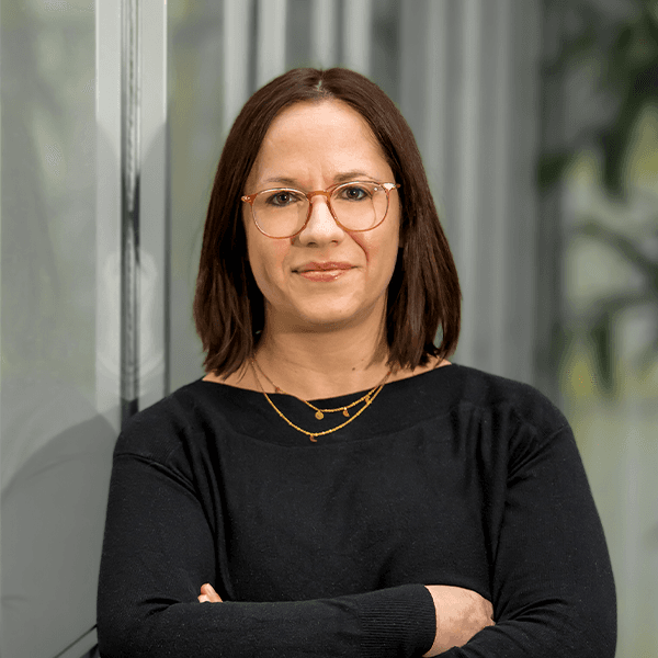 A person with shoulder-length dark hair and glasses stands with arms crossed, wearing a black sweater and gold necklaces, against a backdrop of glass and greenery.