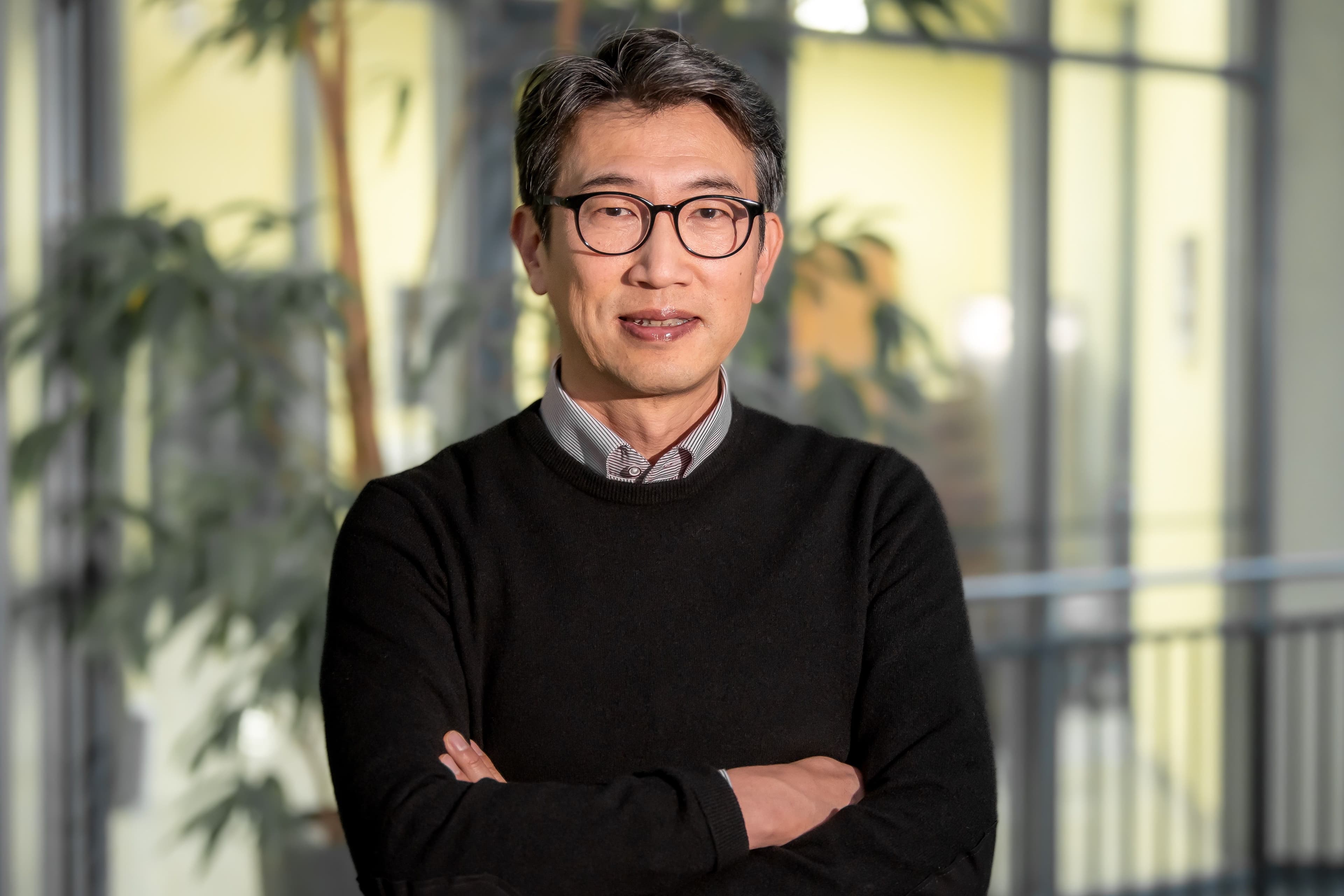 A man wearing glasses and a black sweater stands with his arms crossed in front of a blurred indoor backdrop with plants.