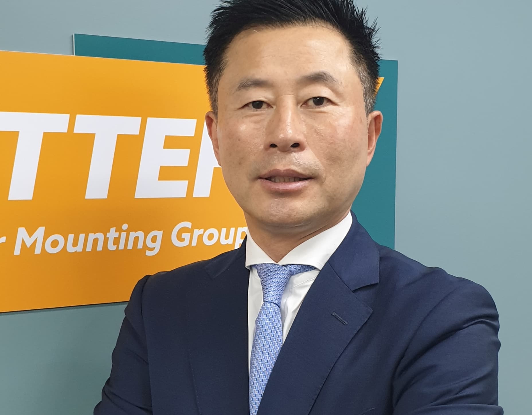 A man in a navy-blue suit and light blue tie stands in front of a sign with partially visible text. He has short black hair and is looking directly at the camera with his arms crossed.