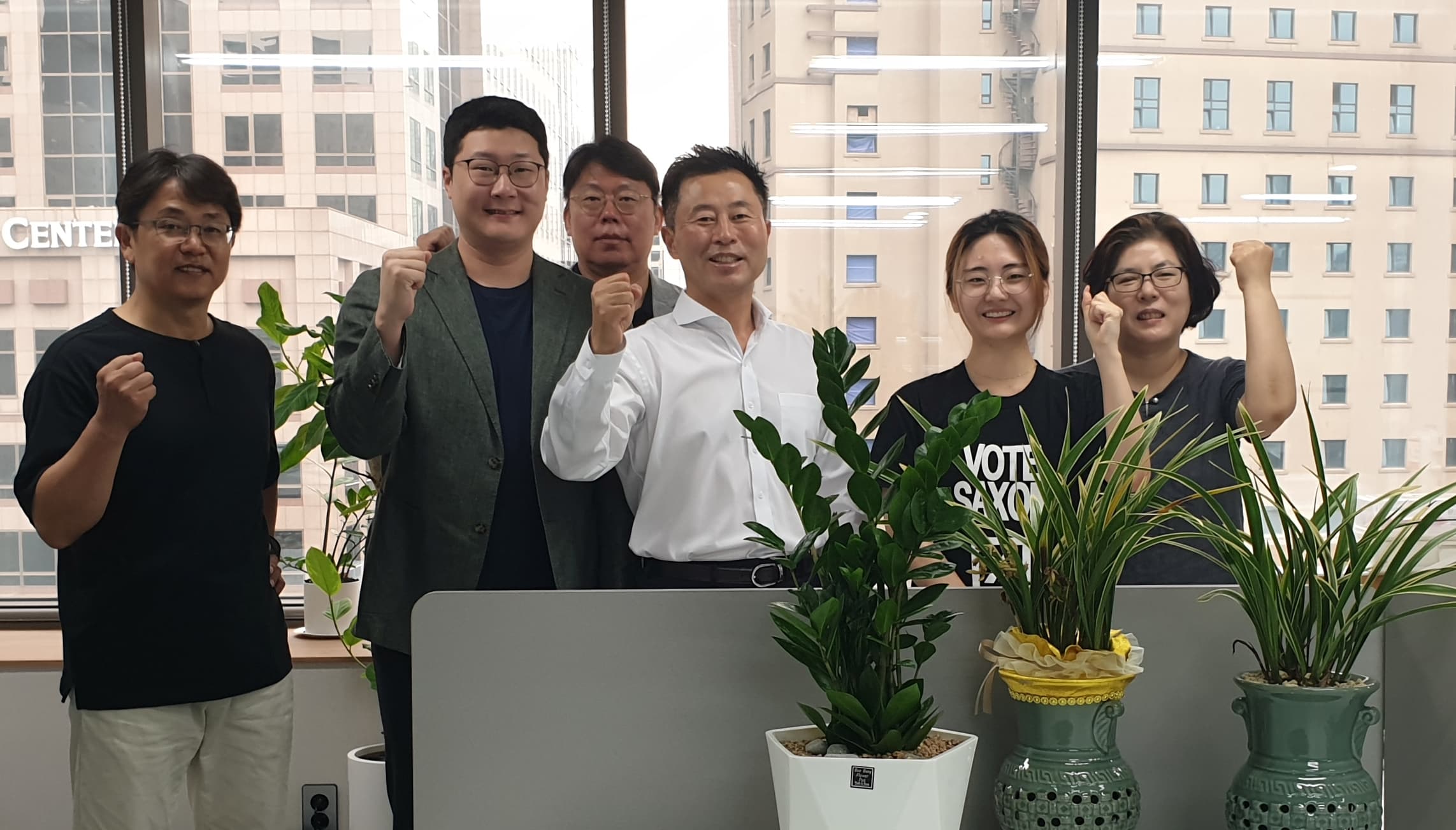 A group of six people standing indoors with city buildings visible through the windows behind them. They are smiling and making fist gestures, with potted plants in the foreground.