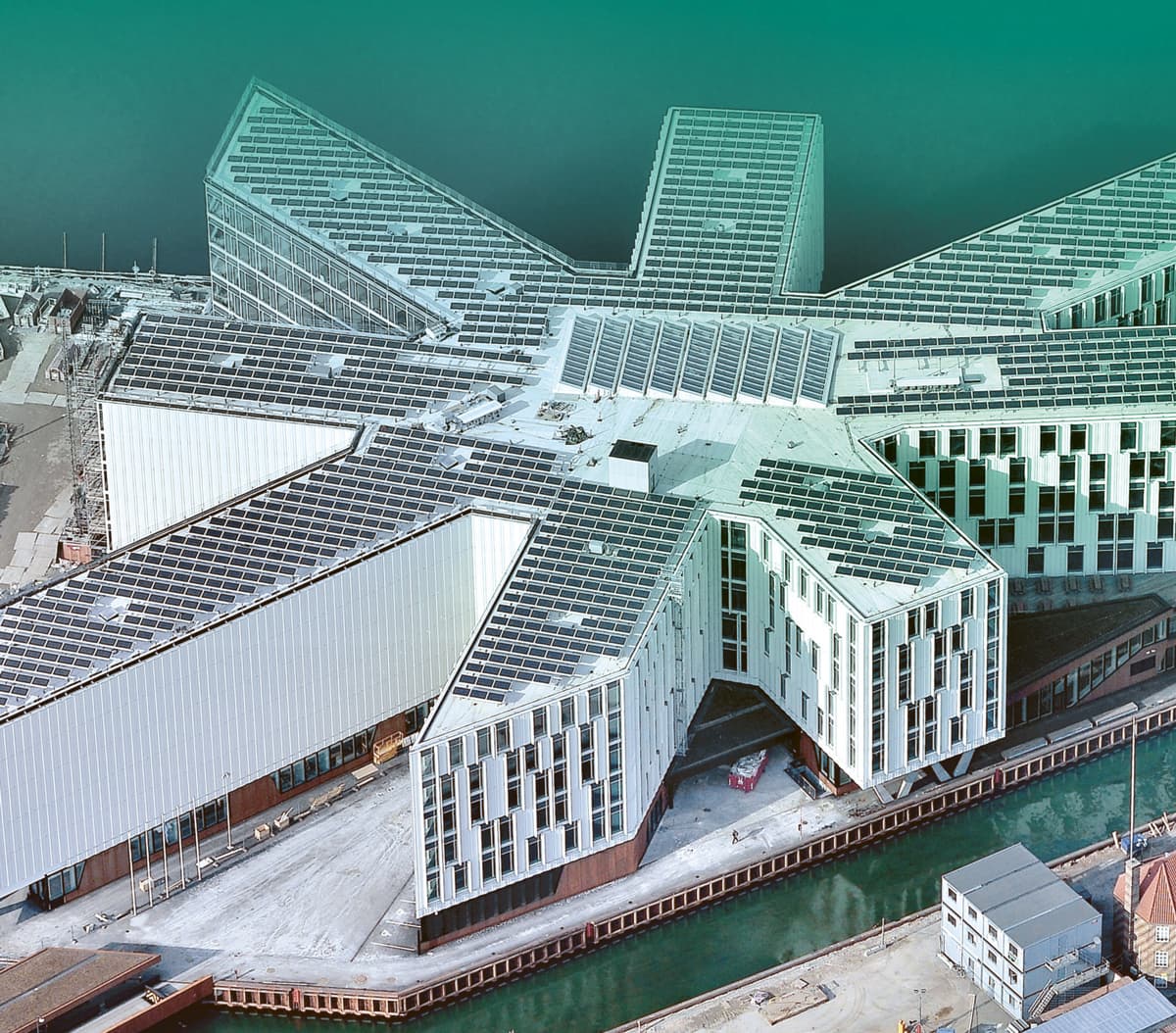 Aerial view of a large, modern, star-shaped building with multiple wings, situated next to a waterway. The building features numerous windows and solar panels on the roof.