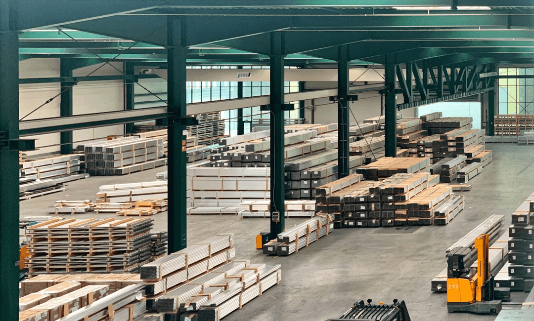 An aerial view of a spacious warehouse with organized stacks of metal, wood, and other materials. Forklifts and shelving units can be seen throughout the facility.