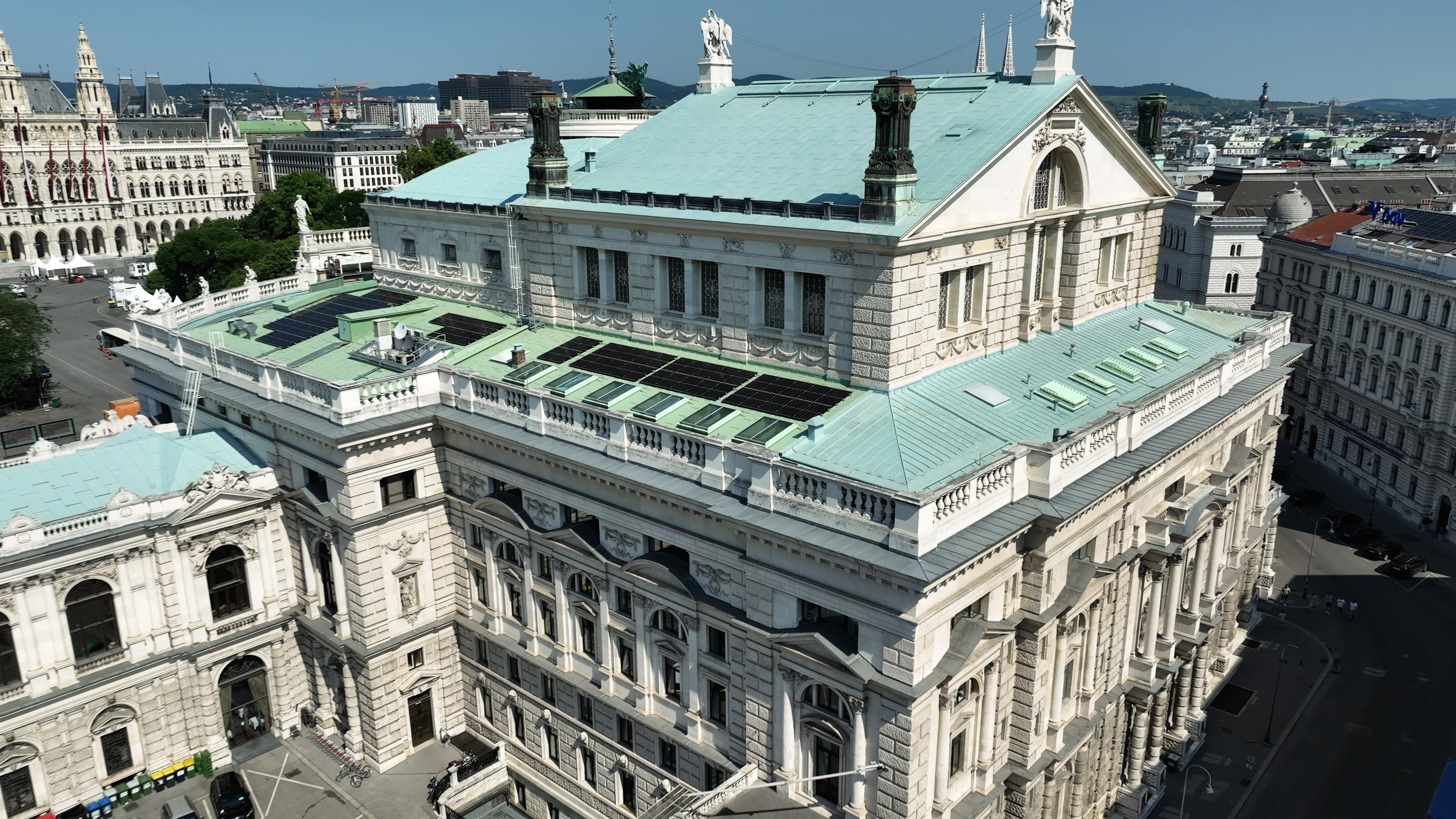 Aerial view of a large, historic building with a green roof and solar panels, situated in a city with other architectural structures visible in the background.