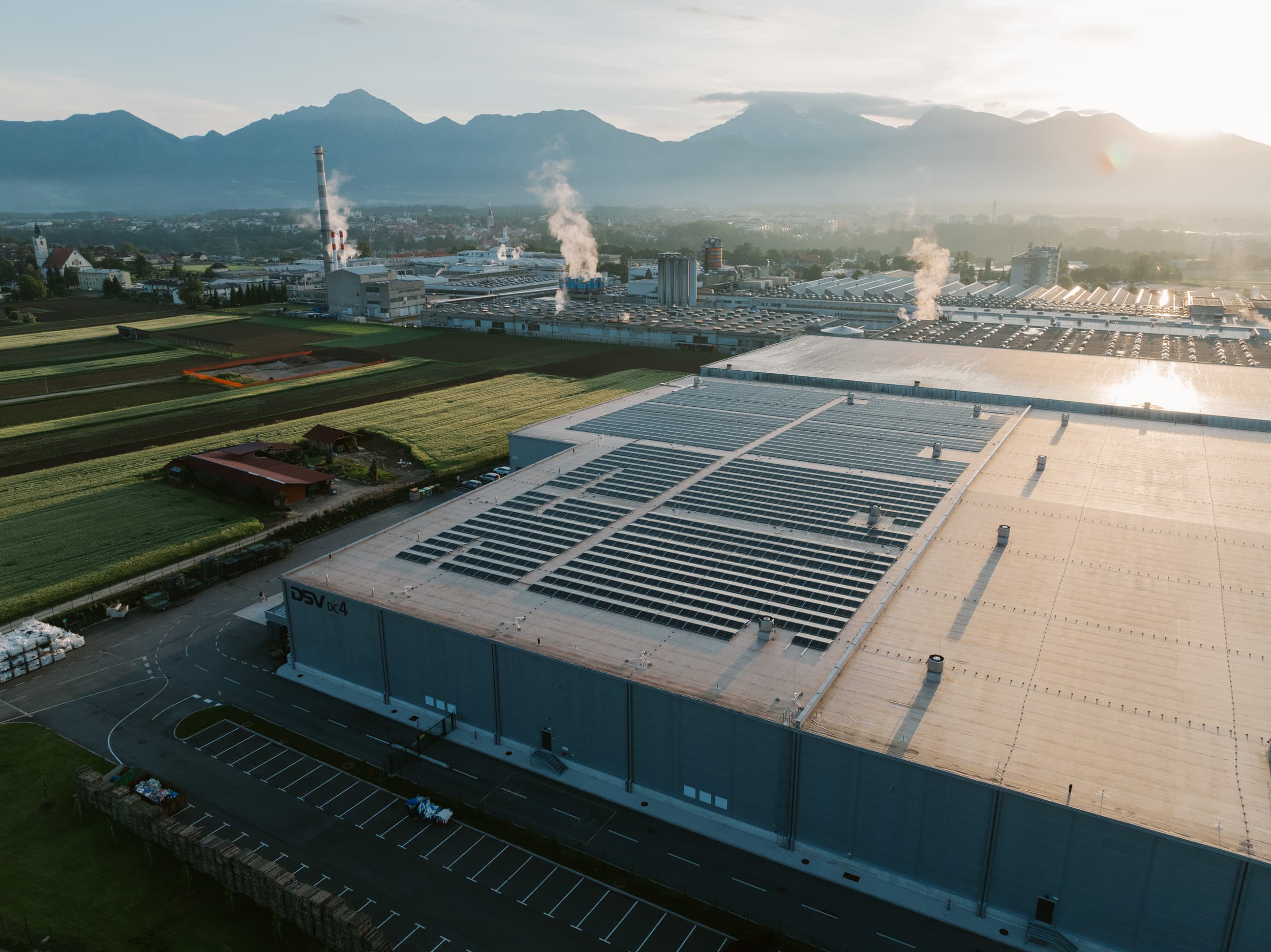 Aerial view of an industrial complex with large buildings, solar panels on the roof, and steam rising from chimneys, set against a backdrop of mountains at sunrise.